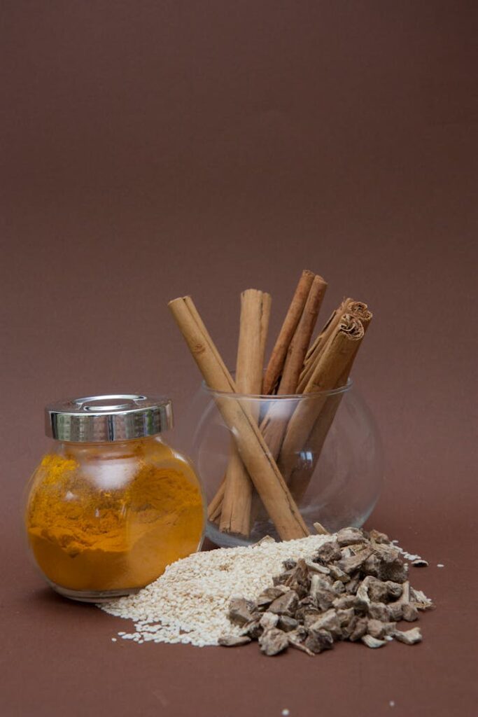 Various spices including turmeric and cinnamon displayed in glass containers with sesame and asafoetida on a brown background.