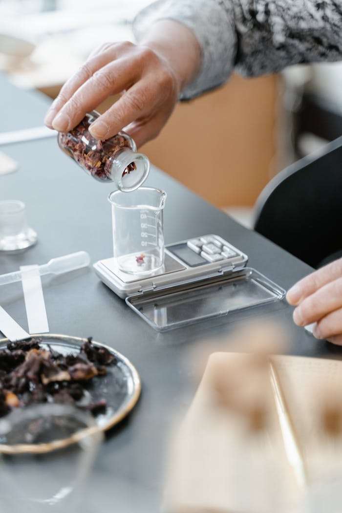 Services A close-up of hands measuring dried flowers into a beaker on a digital scale, demonstrating laboratory aromatherapy preparation.