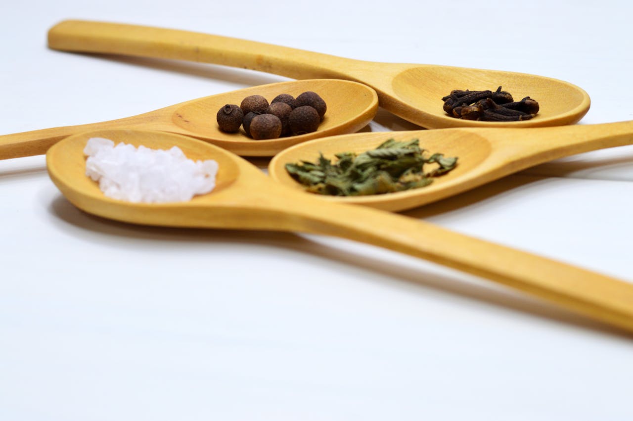About Wooden spoons showcasing salt, cloves, peppercorns, and herbs on a white surface.